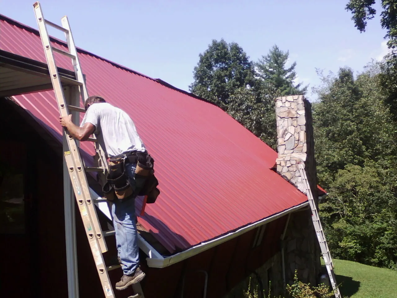 Roofer on a ladder beside a red metal roof and stone chimney — Soffit & Fascia Repair by Roofing Brothers in Mount Victory